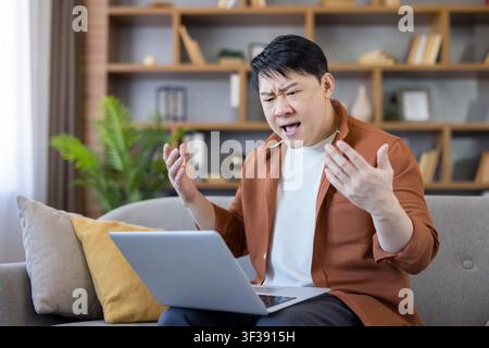Asian man sitting on a sofa and using a laptop, feeling stressed and confused while having a problem with technology, gesturing with hands and yelling in frustration Stock Photo