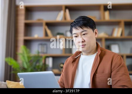 Asian man concentrating on his laptop, working comfortably from his home office, demonstrating remote work, online learning, and focused activity against a modern bookshelf background Stock Photo