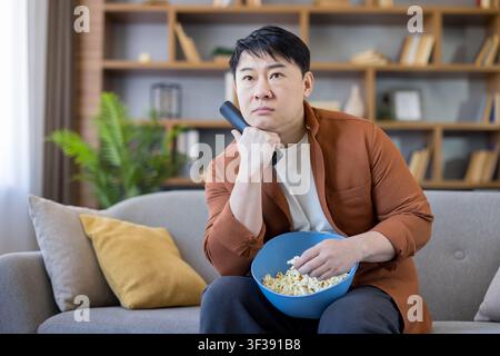 Asian man feeling bored and unimpressed while sitting on a comfortable couch in his living room, holding a remote control and eating fresh popcorn, relaxing at home during leisure time Stock Photo
