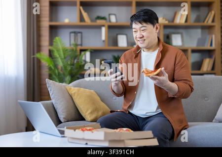 Asian man sitting on a couch at home, enjoying a slice of pizza while smiling and using his smartphone, with a laptop and pizza boxes on the coffee table Stock Photo