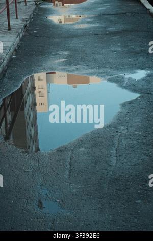 Reflection of a modern building and clear blue sky in a street puddle on cracked, textured asphalt. Stock Photo