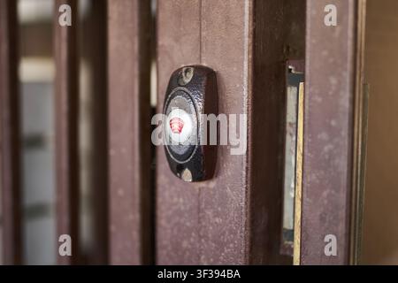 Doorbell Button Mounted On Rusty Metal Gate Closeup Showing Worn Paint Screw Fasteners And Circular Buzzer Housing On Vertical Brown Metal Slats At Exterior Entrance. Stock Photo