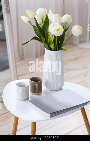 White tulips in vase on table with book or notebook and two cups on table on wooden floor Stock Photo