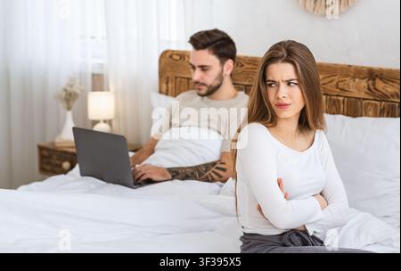 Man works on laptop, woman is offended by bed in bedroom. Stock Photo