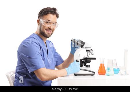 Smiling male scientist in blue scrubs and safety goggles sitting with a microscope Stock Photo