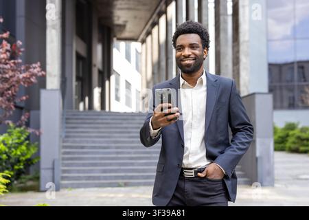 Happy young entrepreneur looking away, holding mobile phone with a broad smile, feeling successful and optimistic while standing outdoors in front of a modern corporate complex Stock Photo