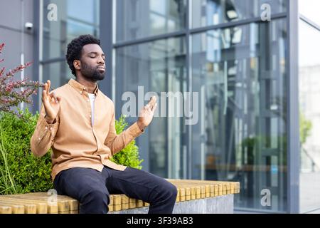 Young man practicing meditation with closed eyes and performing a mudra gesture, finding inner peace, focus, and stress relief during a break outdoors near an office building Stock Photo