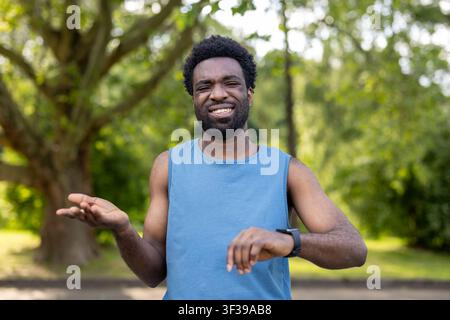 Young man standing outdoors in a park, checking his smartwatch with a frustrated and exasperated expression, indicating potential delays or missed training goals Stock Photo