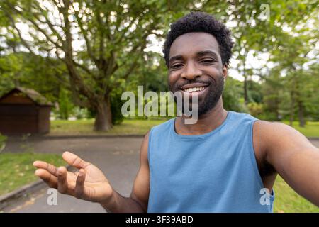 Happy young adult man in a blue tank top making a selfie outdoors, smiling while extending his hand in an inviting or welcoming gesture, enjoying leisure time Stock Photo