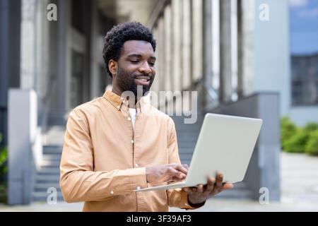 Person smiling and working remotely on a glowing laptop, standing outdoors in an urban environment with a modern building facade, showcasing mobility and connectivity Stock Photo