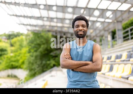 Young man with an athletic build smiling and crossing arms, standing confidently at an outdoor stadium venue, showcasing fitness and determination Stock Photo