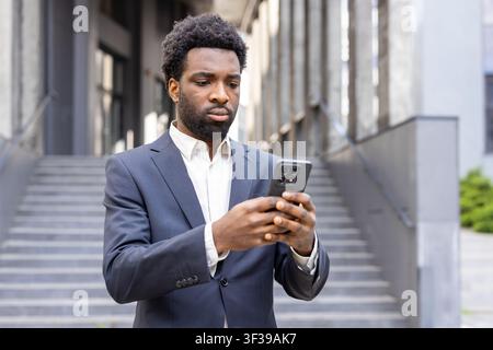 Young businessman engaged with his smartphone while standing outdoors in a city setting, reflecting modern communication and connectivity for professional use Stock Photo