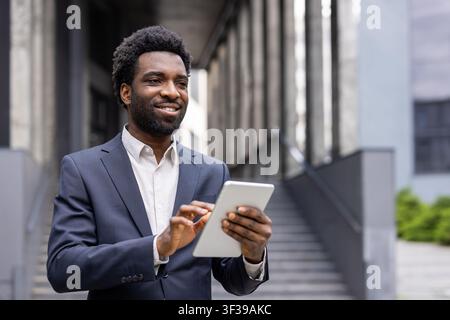 Young businessman standing confidently outside a modern office building, smiling while using a digital tablet, representing success, technology, and professional communication Stock Photo