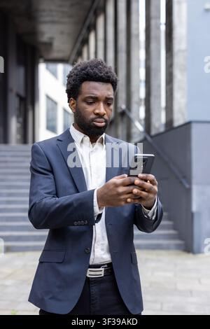 Young businessman standing outdoors, wearing a suit and interacting with his smartphone, actively engaging with mobile technology and digital communication in an urban environment Stock Photo