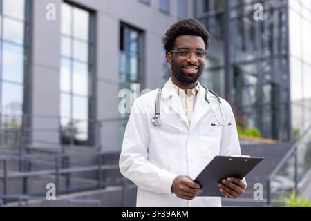 Young african american doctor wearing a lab coat and stethoscope. Confidently holding a clipboard and smiling at the camera in front of a modern medical building Stock Photo