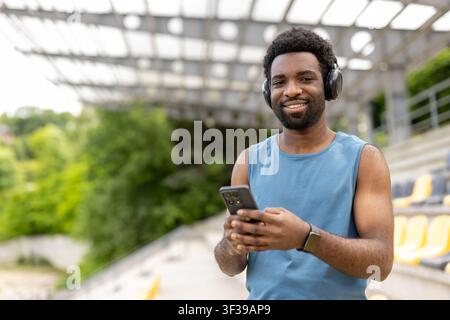 Young man wearing a blue tank top and wireless headphones, holding a smartphone and smiling at the camera outdoors, enjoying music during a wellness break at the stadium Stock Photo
