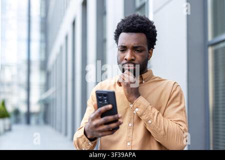 Young businessman in a casual shirt stands outside an office building, staring at his smartphone with a pensive, concerned expression while reading messages and data Stock Photo