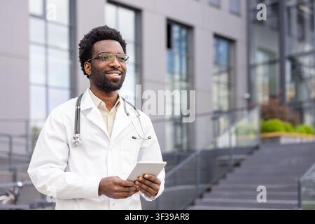Young male medical professional wearing a lab coat and stethoscope, confidently holding a tablet while standing outdoors, representing innovation and technology in healthcare Stock Photo