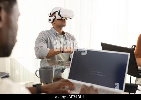 Diverse coworkers watching man wearing white VR headset gesturing over laptops at conference table Stock Photo