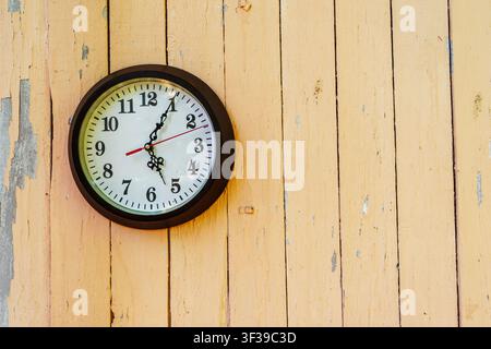 A round wall clock with a white dial and a black frame is mounted on an old wooden wall with peeling yellow paint. The minimalistic composition with plenty of free space is suitable for topics related to time, punctuality, interior design, vintage style and design Stock Photo