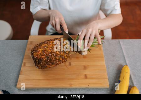 High angle view of woman preparing fresh tropical fruit. Female hands slice pineapple with knife on bamboo cutting board. Healthy eating, food Stock Photo