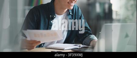 Smiling Young Male Student Holding Papers And Working On Laptop At Home, Study Concept Stock Photo