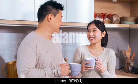 Happy Asian Couple Smiling And Talking While Holding Coffee Mugs In Kitchen, Home Concept Stock Photo