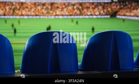 Close up of blue stadium seats in foreground and blurred soccer field with players and fans in background. Empty live sports sports venue tribunes during football match Stock Photo