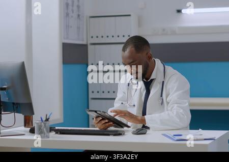 African American therapist doctor sits at a hospital desk with a tablet, reviewing medical documents and typing sickness expertise for treatment. Stock Photo
