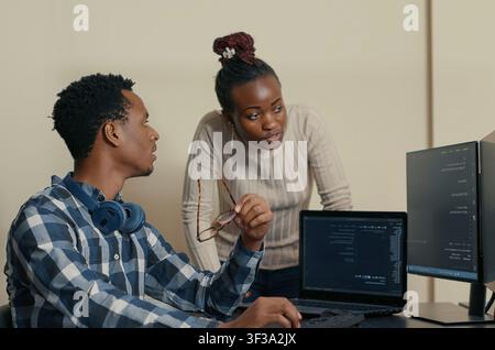 Database developer removes glasses while explaining an algorithm to a colleague, pointing at code on multiple screens during team development. Stock Photo