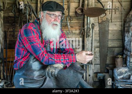 At the historic Alex Matheson Blacksmith’s workshop in the Highland Village Museum | Baile nan Gàidheal in Iona, Nova Scotia, Canada, a blacksmith dressed in period costume demonstrates the craft as practised in the 1920s. Pliers, saws and horseshoes hang on the walls Stock Photo