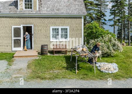 In front of a yellow-painted house at the Highland Village Museum | Baile nan Gàidheal in Iona, Nova Scotia, Canada, a craftsperson in traditional dress is processing freshly shorn sheep’s wool on a wooden frame, whilst another sweeps the steps leading up to the entrance. Bras d’Or Lake can be seen in the background Stock Photo