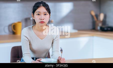 Worried Young Asian Woman Sitting Alone At Kitchen Table With Empty Bowl, Weight Loss Concept Stock Photo
