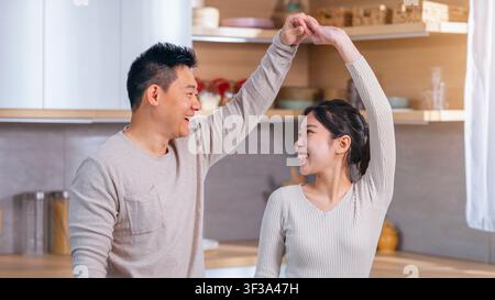 Happy Asian Couple Dancing Together In Modern Kitchen, Smiling And Enjoying Moment Stock Photo