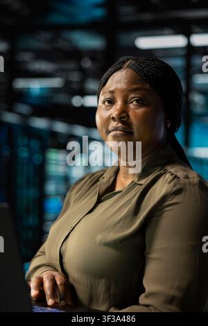 Woman in data center using laptop to troubleshoot backend operations infrastructure rigs. African american server hub worker running software development tools on device, resolving system crashes Stock Photo