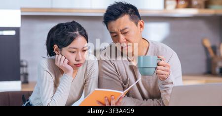 Focused Asian Couple Reviewing Documents In Modern Kitchen, Serious Discussion Stock Photo