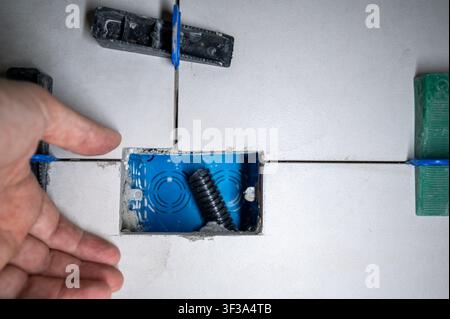 Hand placing ceramic tile around a blue electrical box, using a tile leveling system to ensure perfectly even surfaces during house renovation and con Stock Photo
