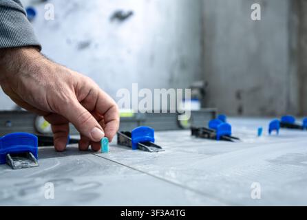 Hand of a tiler installing new gray ceramic floor tiles on adhesive mortar, using a tile leveling system to ensure a perfectly even and flat surface d Stock Photo