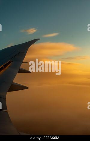 Airplane wing silhouetted against a backdrop of golden sunset clouds, visible through an aircraft window during flight. Stock Photo