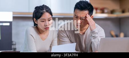 Worried Asian Couple Examining Financial Documents At Home, Concerned Expressions Stock Photo