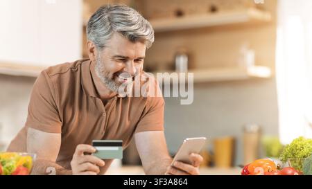 Happy Senior Caucasian Man Using Smartphone And Credit Card In Kitchen, Online Shopping Concept Stock Photo