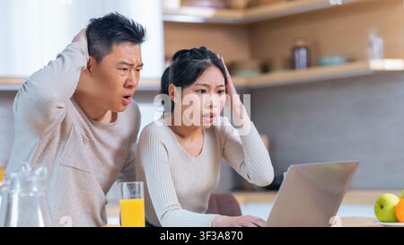 Worried Asian Couple Looking At Laptop In Kitchen, Stressful Situation Stock Photo
