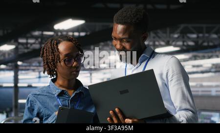 Data center system administrators using laptop to review AI LLM visualizations. African american IT specialists use device to analyze neural networks outputs, ensuring stable gear operations Stock Photo