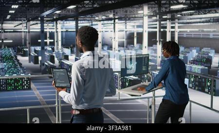 Data center programmers on industrial platform checking lines of code, collaborating on tasks. Server hub IT specialists visualizing large language model architecture using tablet and PC Stock Photo
