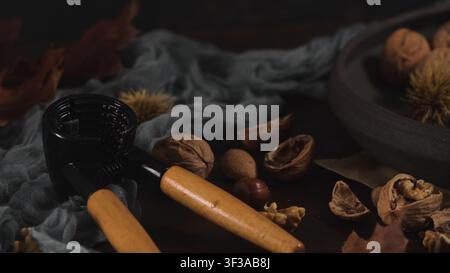 A dark moody close-up highlights a black metal nutcracker with wooden handles alongside scattered walnuts, shells, and spiky chestnut burrs on a drape Stock Photo
