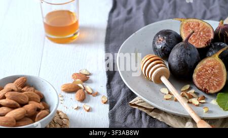Fresh whole and sliced figs are arranged on a ceramic plate next to a wooden honey dipper and almonds, illuminated by bright natural light on a rustic Stock Photo