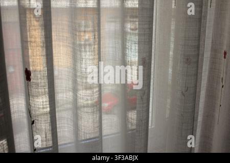 A medium shot of a sheer white tulle curtain featuring delicate embroidered flowers, hanging in front of a window with a softly blurred background sho Stock Photo
