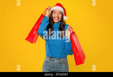 Happy woman in Santa hat holding shopping bags, yellow background Stock Photo