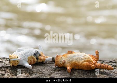 Chiang Mai, Thailand - 13 July 2026 : Calico and orange tabby cat figurines sleeping side by side on a mossy tree root beside a waterfall Stock Photo