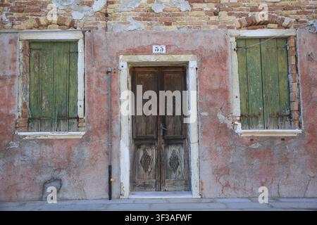 Two windows and a door in an old, weathered red brick facade, windows with green shutters, colorful houses on the island of Burano, Venice, Veneto, I Stock Photo